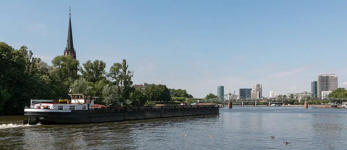 A barge glides along the calm River Main past leafy banks and a church spire